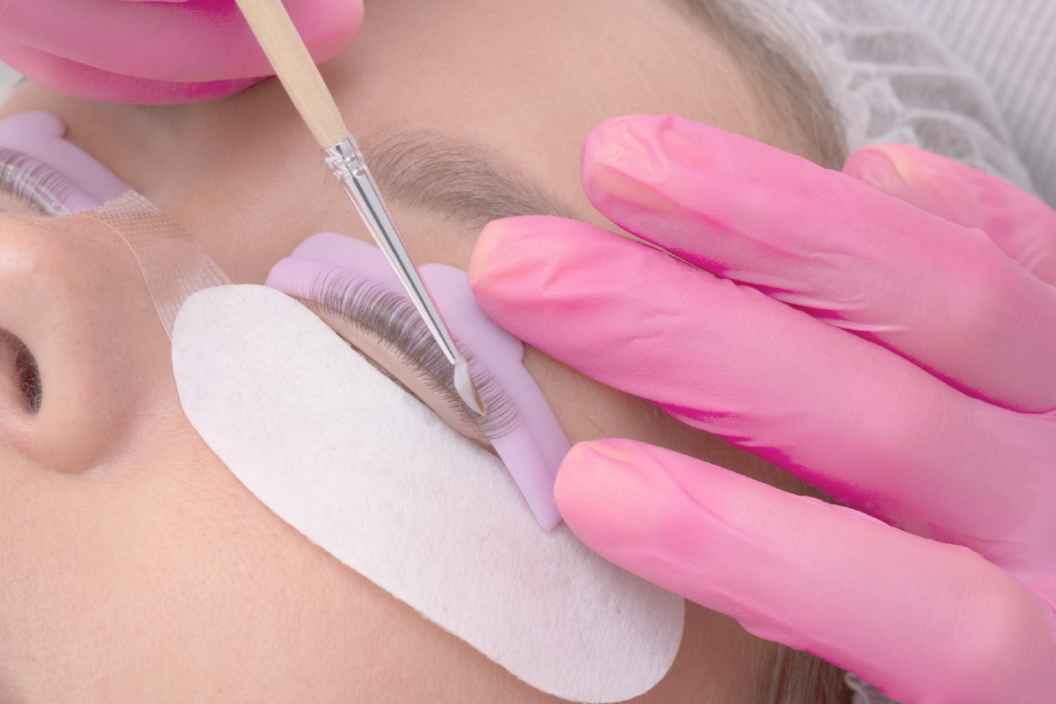 Close-up of eyelash extension glue being applied to natural lashes with precision, using eye pads and pink gloves.